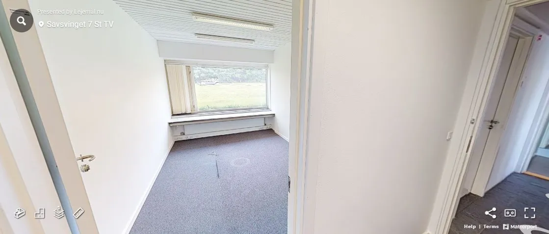 En tom kontorlokal i Savsvinget 7 med lyse loftslampe, hvidt panel og gulvbelægning og et stort vindue med udsigt til en