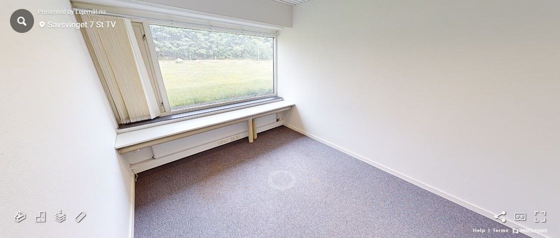 Et tomt værelse med hvide vægge og et gulvtæppe. Under vinduet er der en bænk med radiator og en stikkontakt.