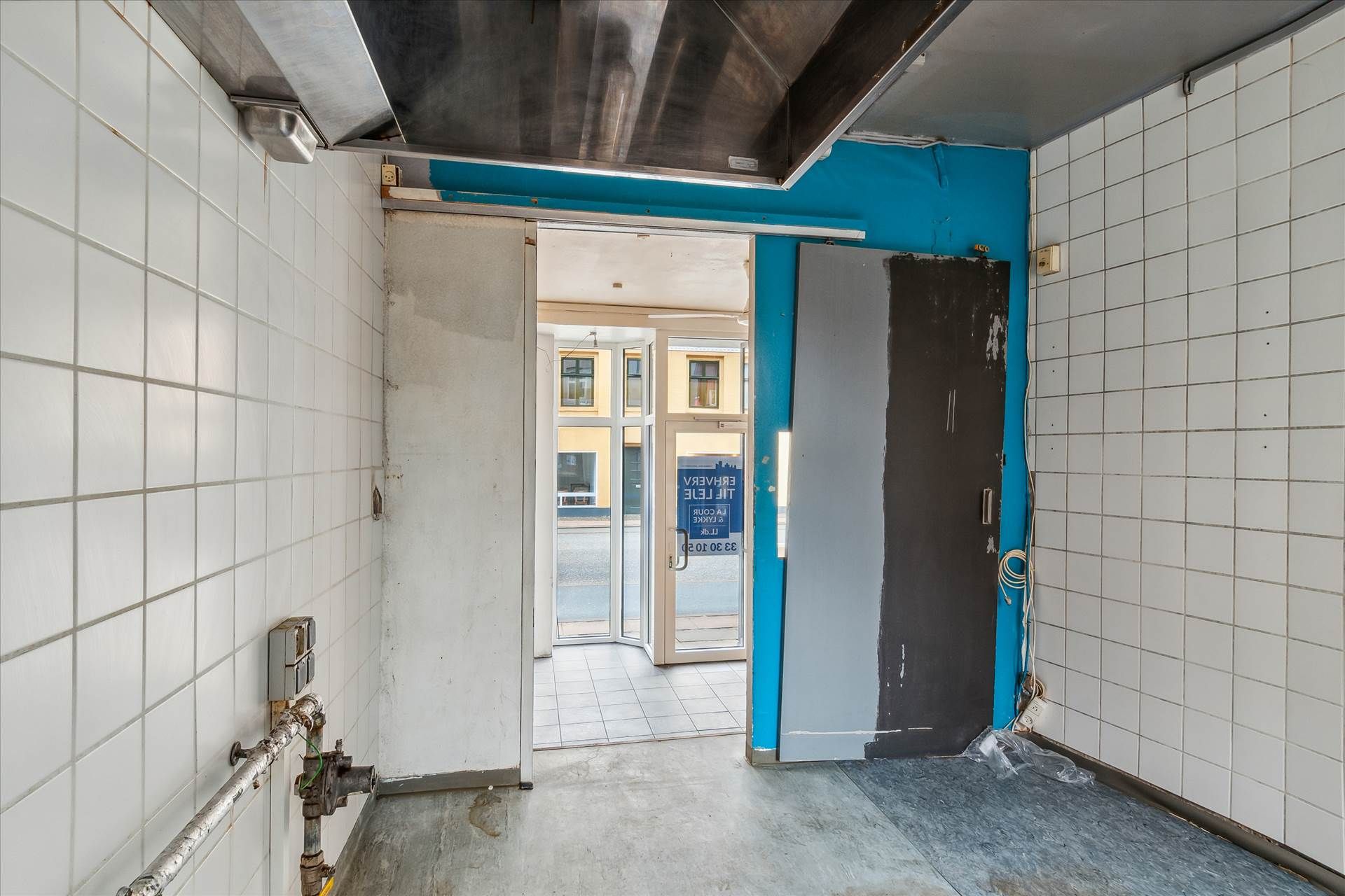 En ubenyttet handelslokal med hvide kakeloverflader og glasdøre samt en skraldkurv og en elektrisk slukkontakt