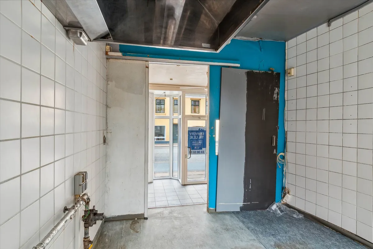 En ubenyttet handelslokal med hvide kakeloverflader og glasdøre samt en skraldkurv og en elektrisk slukkontakt