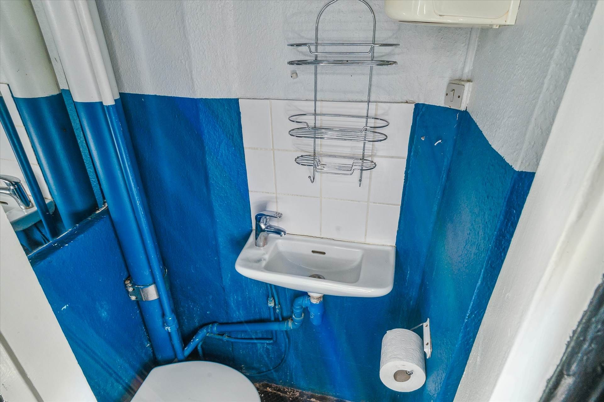 Lille badeværelse med hvid og blå væg og et toilet, et hængende toiletpapir, en hængende metalholder og en håndvask med
