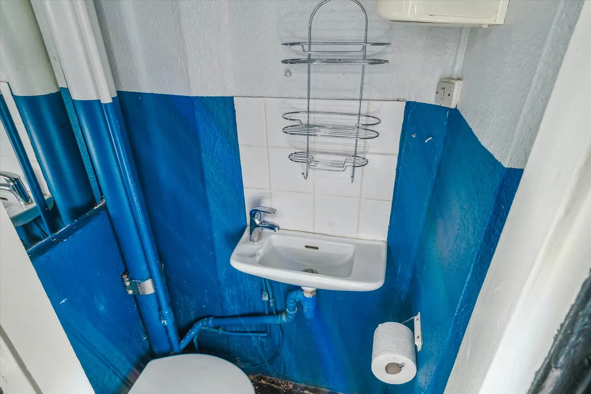 Lille badeværelse med hvid og blå væg og et toilet, et hængende toiletpapir, en hængende metalholder og en håndvask med