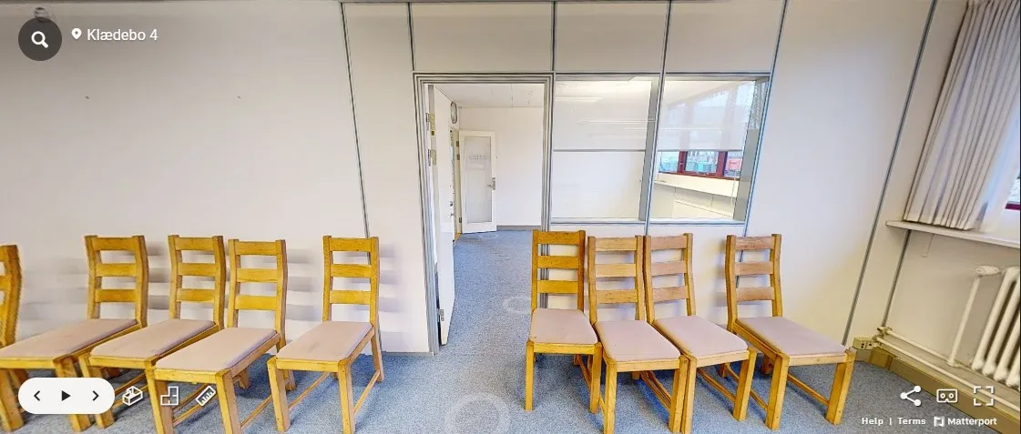 Rum med to rækker lysebrune stole, der er arrangeret med højdepunkter mod en hvid væg. Rummet er tomt og har gulvbelægning