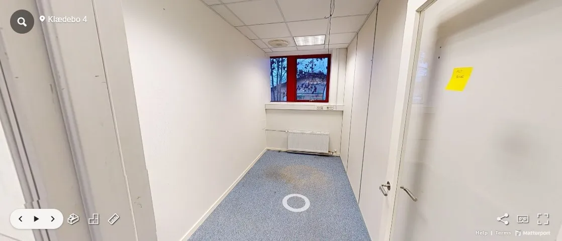 Et lyst og rent kontor med lysegule vægge, et vindue og en hvid dør. Gulvet er blåt og har et spor på det. Der er et gult