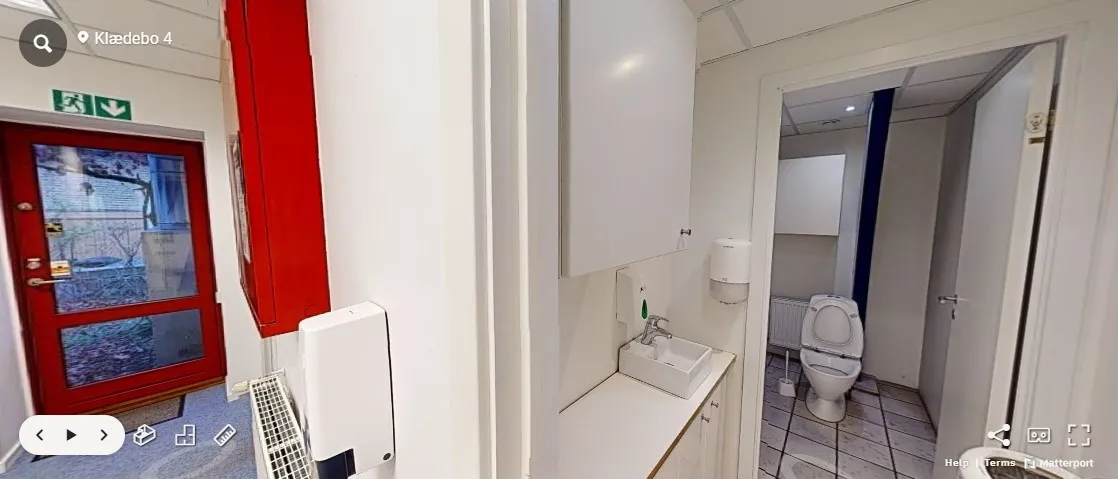 Kontor med hvid og rød væg og rødt dørblad, toilet med toiletpapir og håndvask med vandhane, hvidt loft og gulv, åben dør og