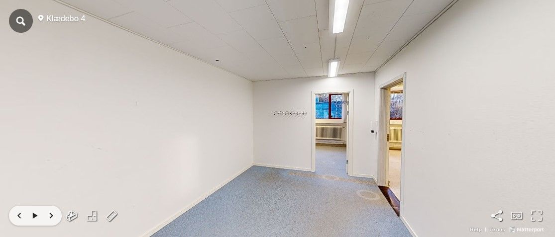 Foto af et tomt kontor med hvide vægge, blå gulv og åbne døre. Der er skrift på væggen og en radiator mod loftet.