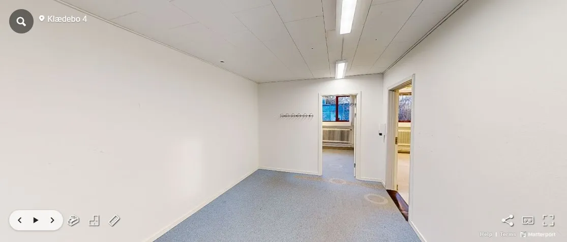 Foto af et tomt kontor med hvide vægge, blå gulv og åbne døre. Der er skrift på væggen og en radiator mod loftet.