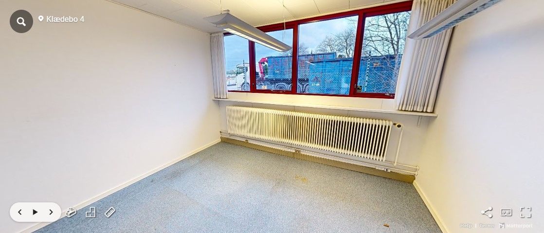 Et tomt kontorlokale med hvide vægge, hvid gulvbelægning, radiatorer og trækruderede vinduer. Et tungt køretøj er synligt