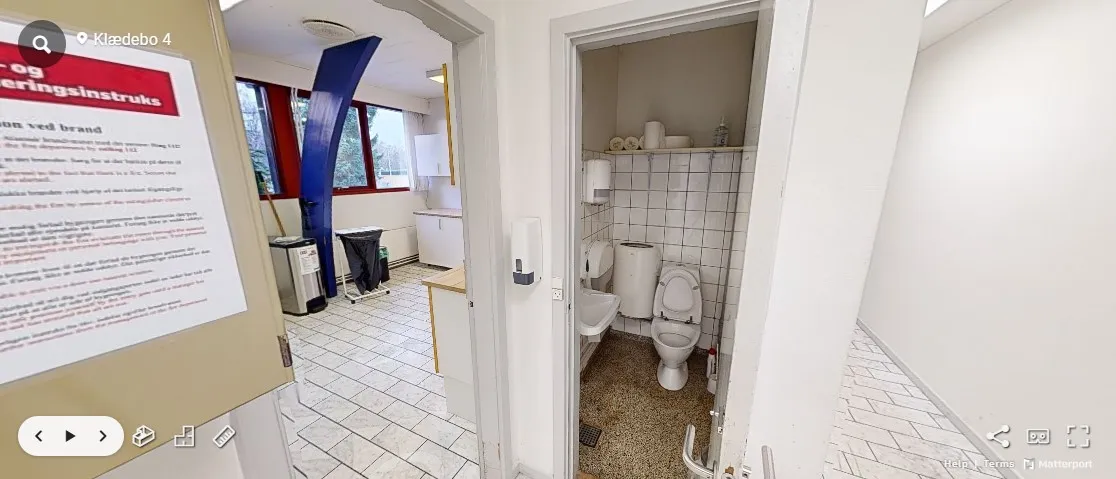 Toiletrum med toilet, håndvaskeskål og to toiletpapirholder. Der er et rengøringsrum med skraldspande og rengøringsudstyr.