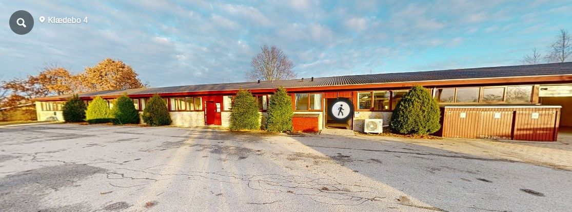 Kontorbyggeri med røde døre og grønne buske i forgrunden. Stående skilt med et gående menneske. Foran bygningen er der en