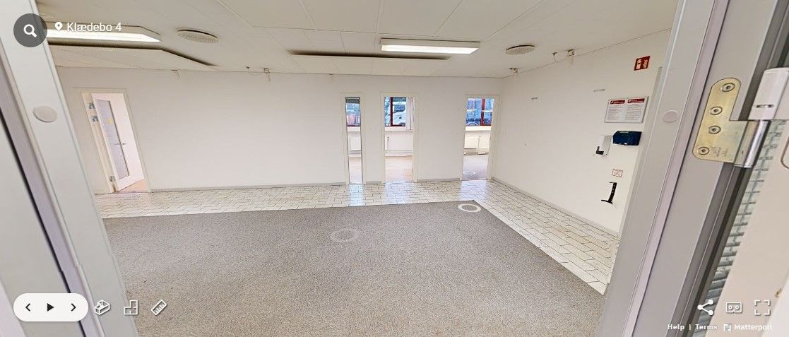 Tomt rum med hvide vægge, lyse gulvbelægning og to indgange, én med åben dør og en anden med lukket dør.