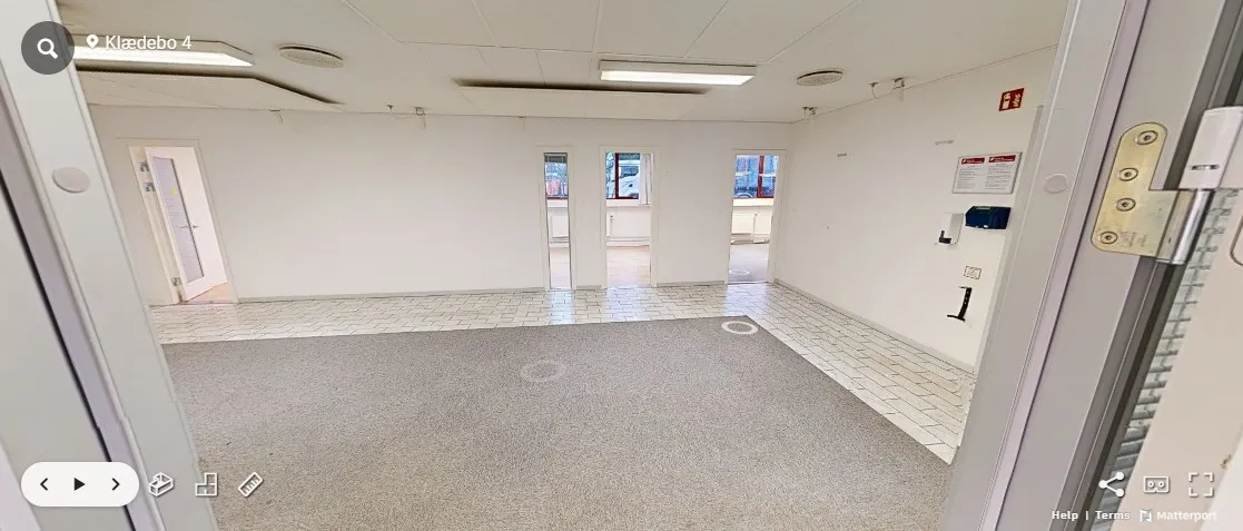 Tomt rum med hvide vægge, lyse gulvbelægning og to indgange, én med åben dør og en anden med lukket dør.