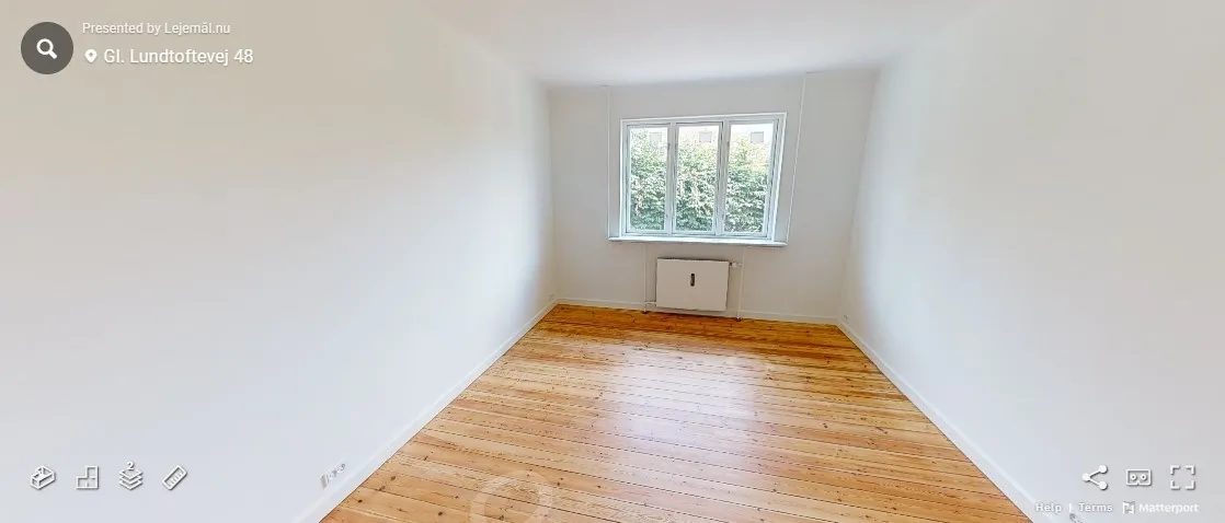 Ledig og nyrenoveret værelse med hårdt trægulv, hvidt vægmaleri og en hvid radiator mod væggen. Der er et slankt vindue med