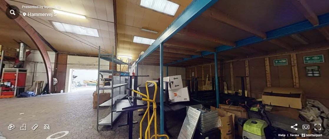 Lager med lagerhylder og opbevaringsbokse. Der er flere stålreoler og et gult stativ. Der er flere papæsker og kasser. Der