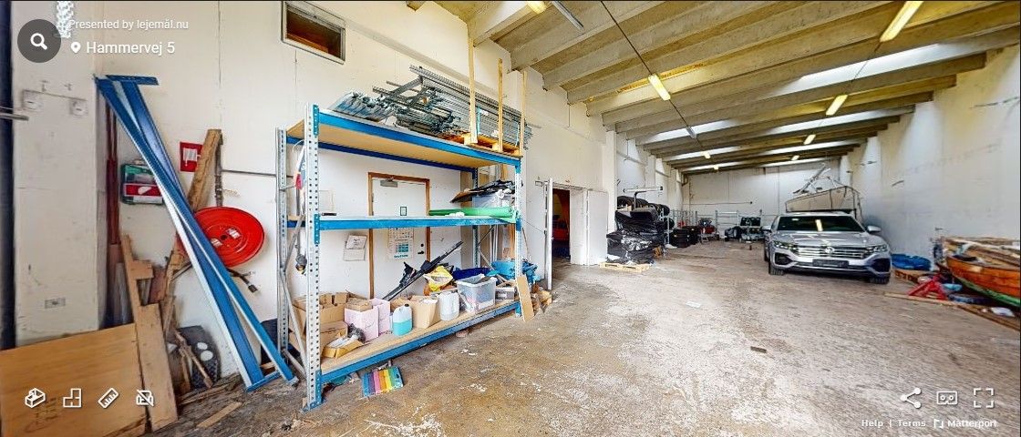 En meget stor værkstedslokal med store, lyse lofter og en alu port i midten af lokalet. Et lager er fyldt med forskellige