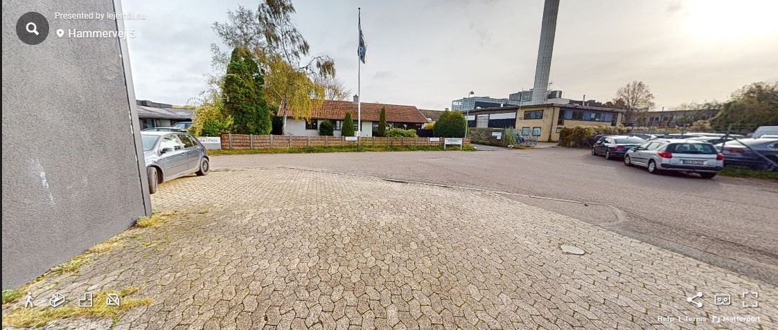 En bilparkeringsplads med flere parkerede biler, slanke træer, et højt flag og en husrække i baggrunden.