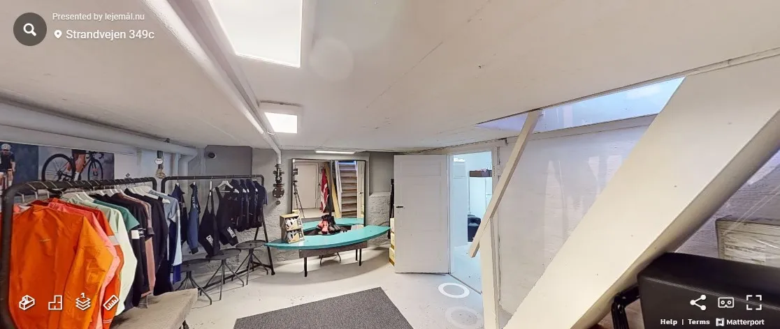 En stor stue med tøj på stående reoler og en grøn skrivebord på gulvet. Der er også en række cykler og tøj i loftet.