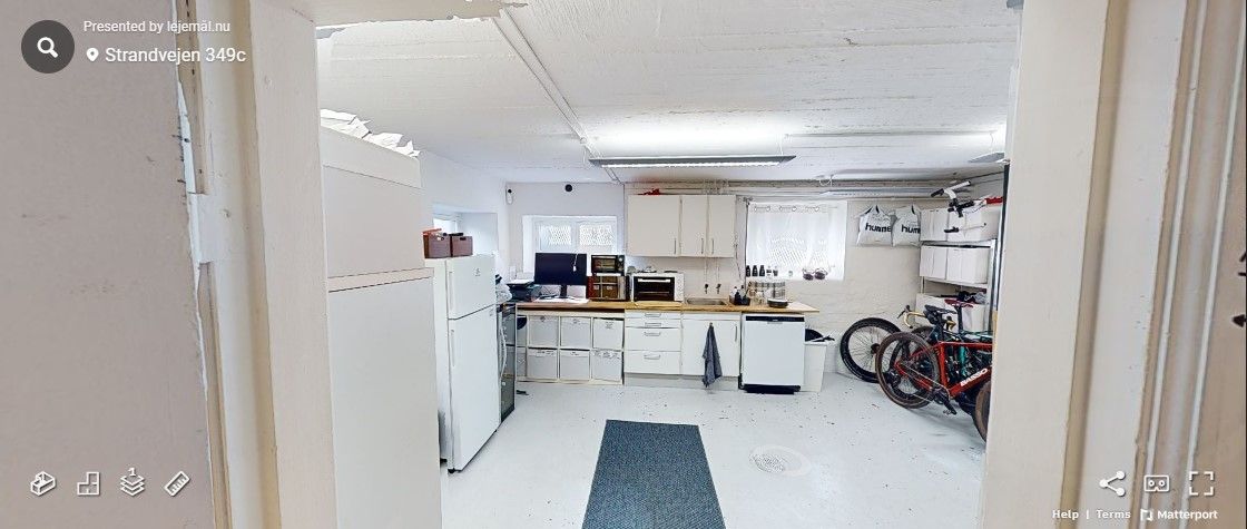 Et køkken med hvide vægge og loft, hvor der står en hvid køleskab og flere apparater. Der er et gult gulvtæppe og en række