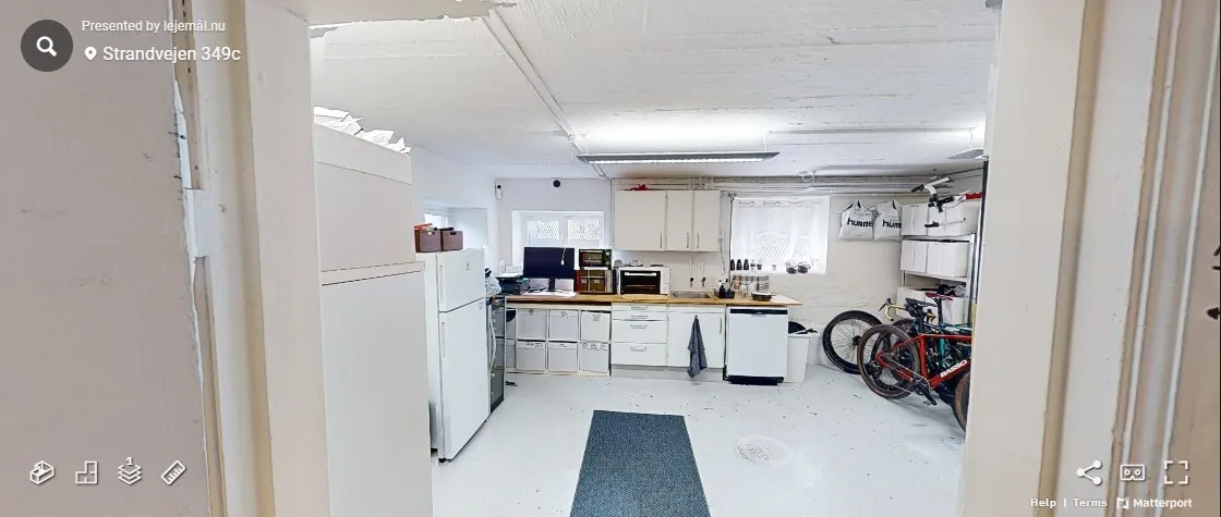 Et køkken med hvide vægge og loft, hvor der står en hvid køleskab og flere apparater. Der er et gult gulvtæppe og en række