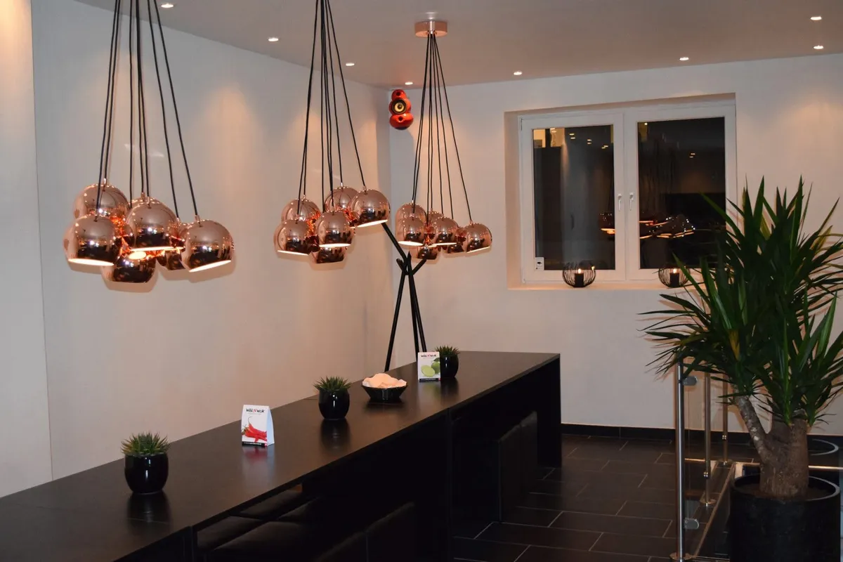 Et moderne interiør med lange borde og røde lysekroner med kugleformede lamper samt en stor plante i sort pot.