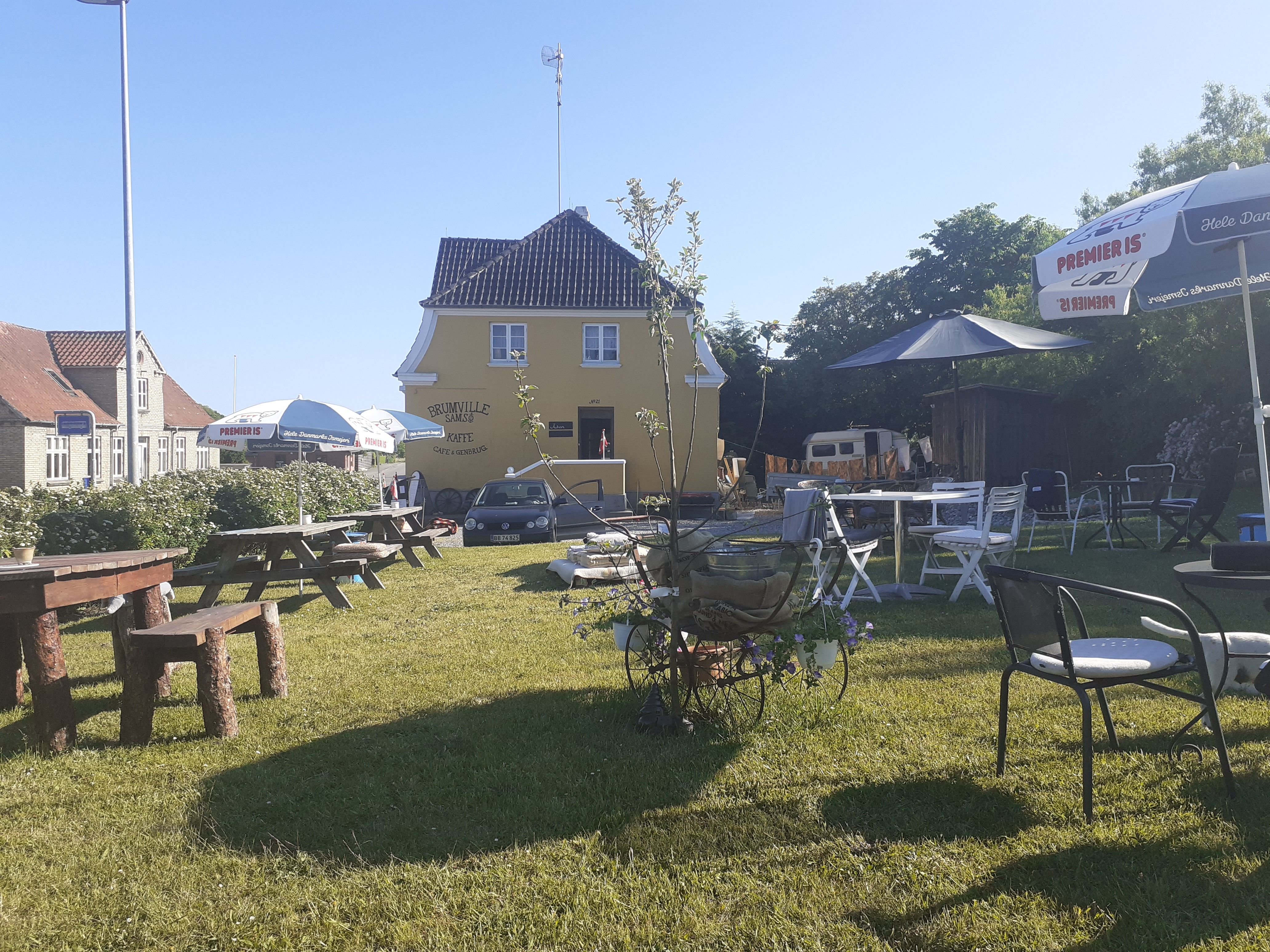 En hyggelig gård med lyse gule vægge, hvor der er flere bordsæt og parasoller udenfor. Gården er omgivet af grønne planter