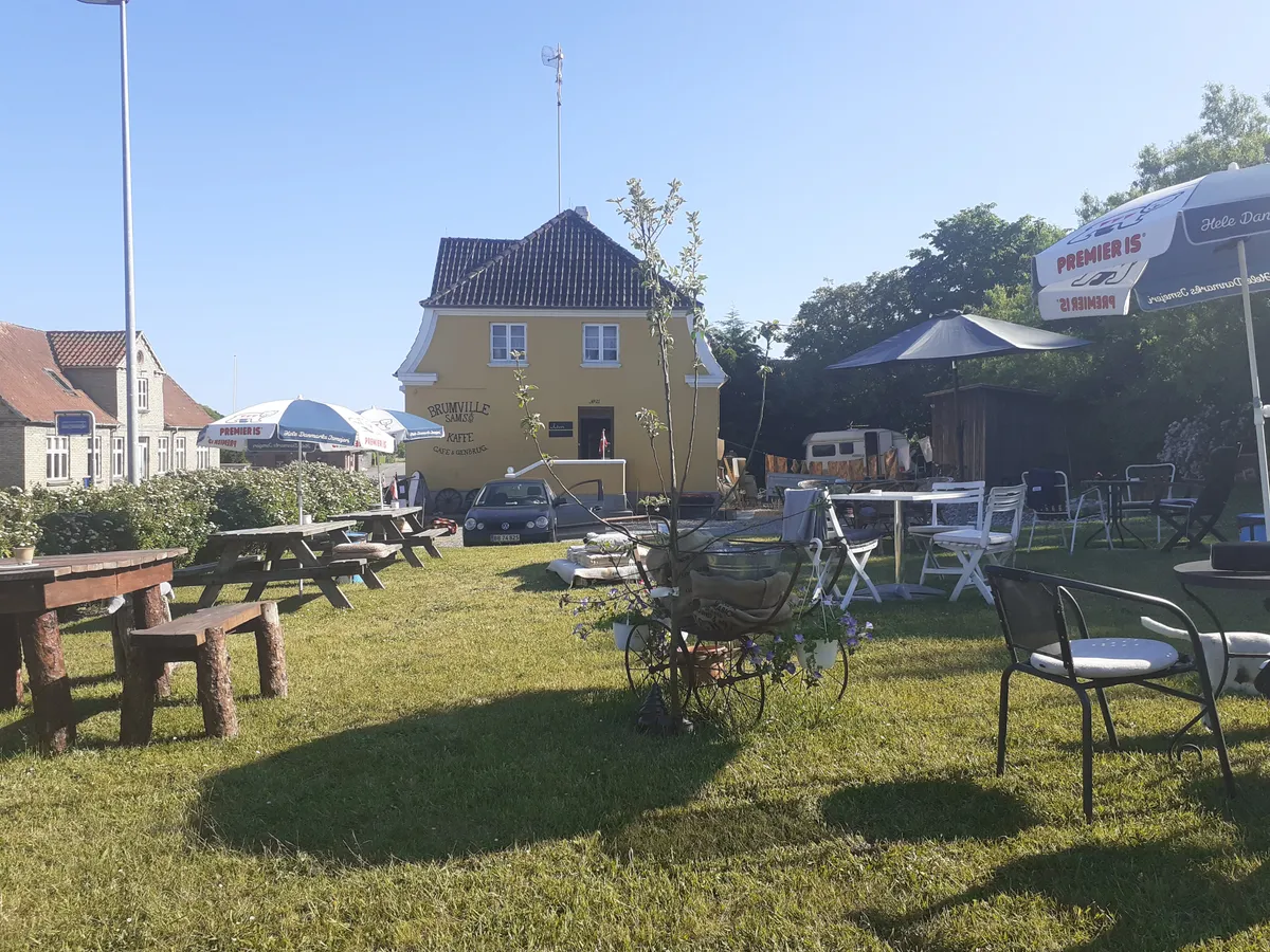 En hyggelig gård med lyse gule vægge, hvor der er flere bordsæt og parasoller udenfor. Gården er omgivet af grønne planter