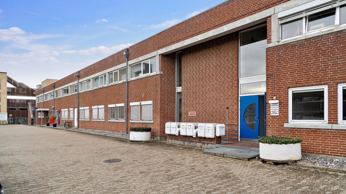 En stor bygning med røde mursten og hvide vindueskarm. Der er en hvid kasse foran døren, der er blå. Der er en lille stier