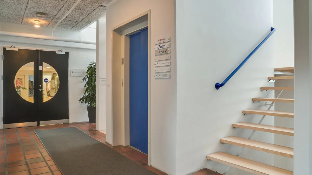 Kontorbygning med lyse møbler og trappe med håndtag, indgangsdøre og gulvbelægning af røde og gule teglsten samt hvid væg