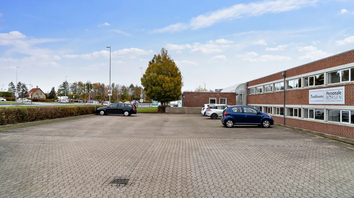 Parkeringsplads med flere parkerede biler foran et kontorhuse med skilte med teksten Personlig Borsen og TeamVikaren