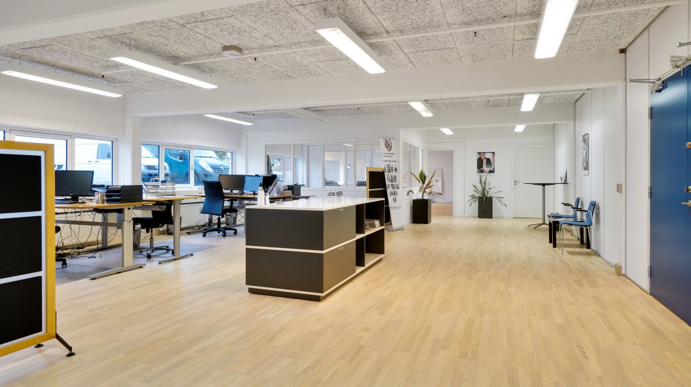 Moderne kontor med lyse trægulve, computerborde, skriveborde og to skyggeslagte hjørner med planter og billeder