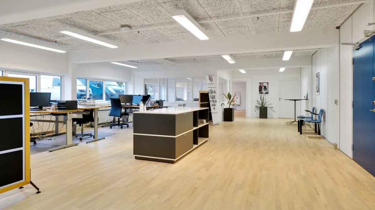 Moderne kontor med lyse trægulve, computerborde, skriveborde og to skyggeslagte hjørner med planter og billeder