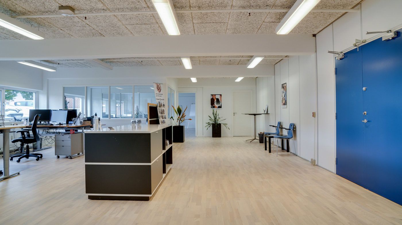 Kontorlokaler med lyse gulve, højt loft og store vinduer. To skriveborde med computere og en sofa med to stole. Planter og