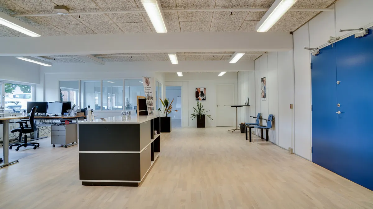 Kontorlokaler med lyse gulve, højt loft og store vinduer. To skriveborde med computere og en sofa med to stole. Planter og