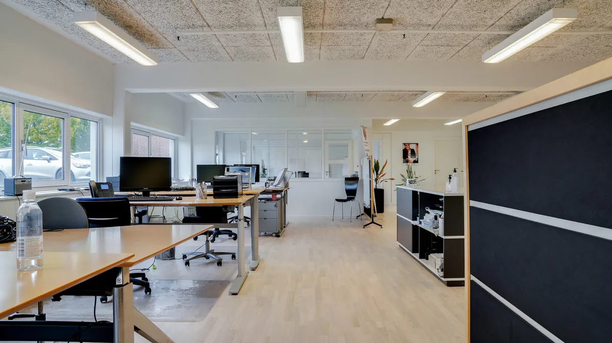 Kontorlokaler med skriveborde, stol, computer, telefon, plante, lyskabler og åbne ruder i en stor rummelig stue med en