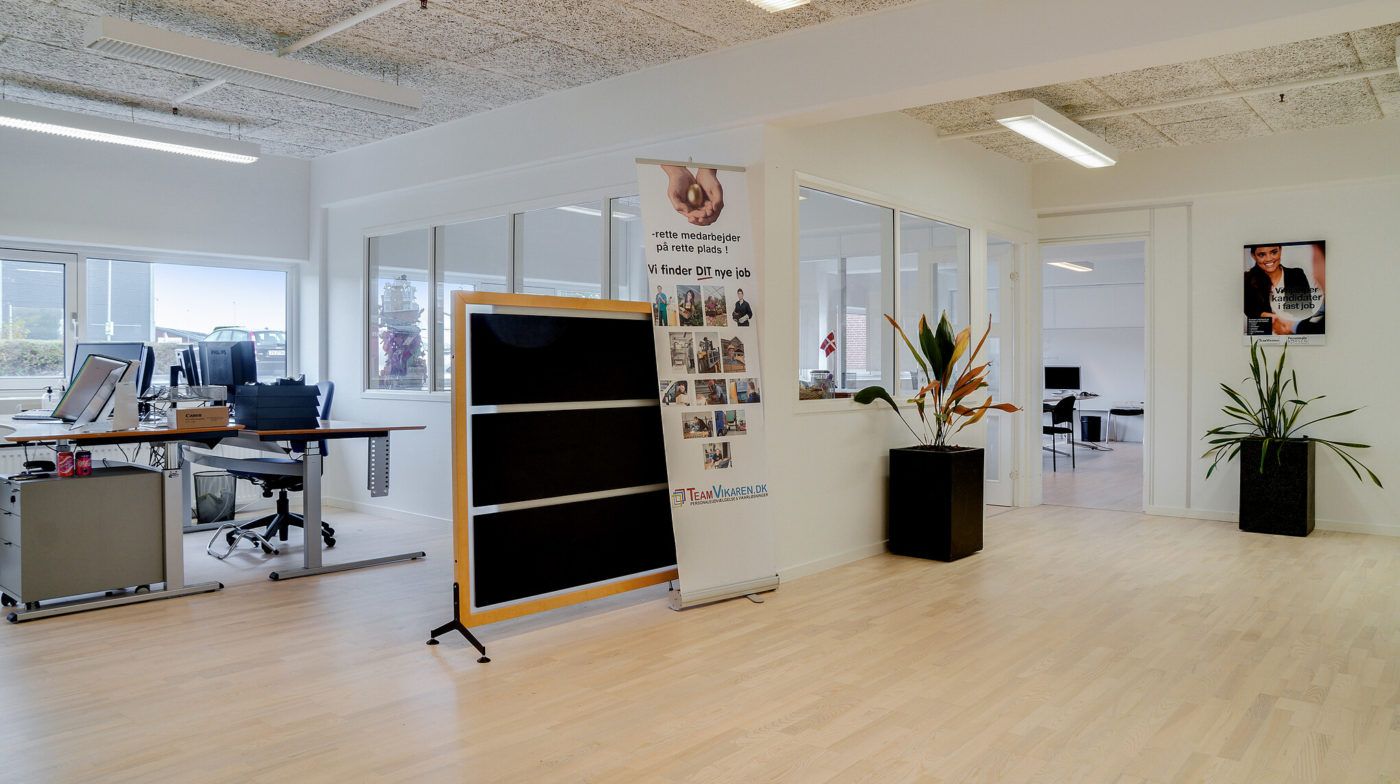 Kontorområde med skriveborde og stole, to plantepot med planter og reklameplakat med billeder og tekst samt vinduer med