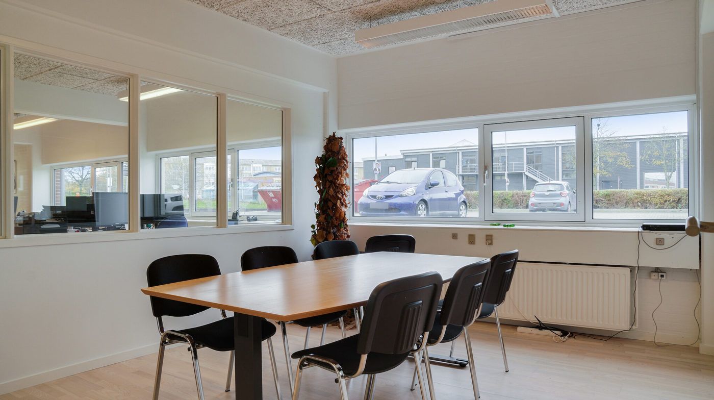 En kontorudlejning med en stor vinduesfront, en træbord og flere stole, et klimaanlæg og et landskabsbillede udenfor