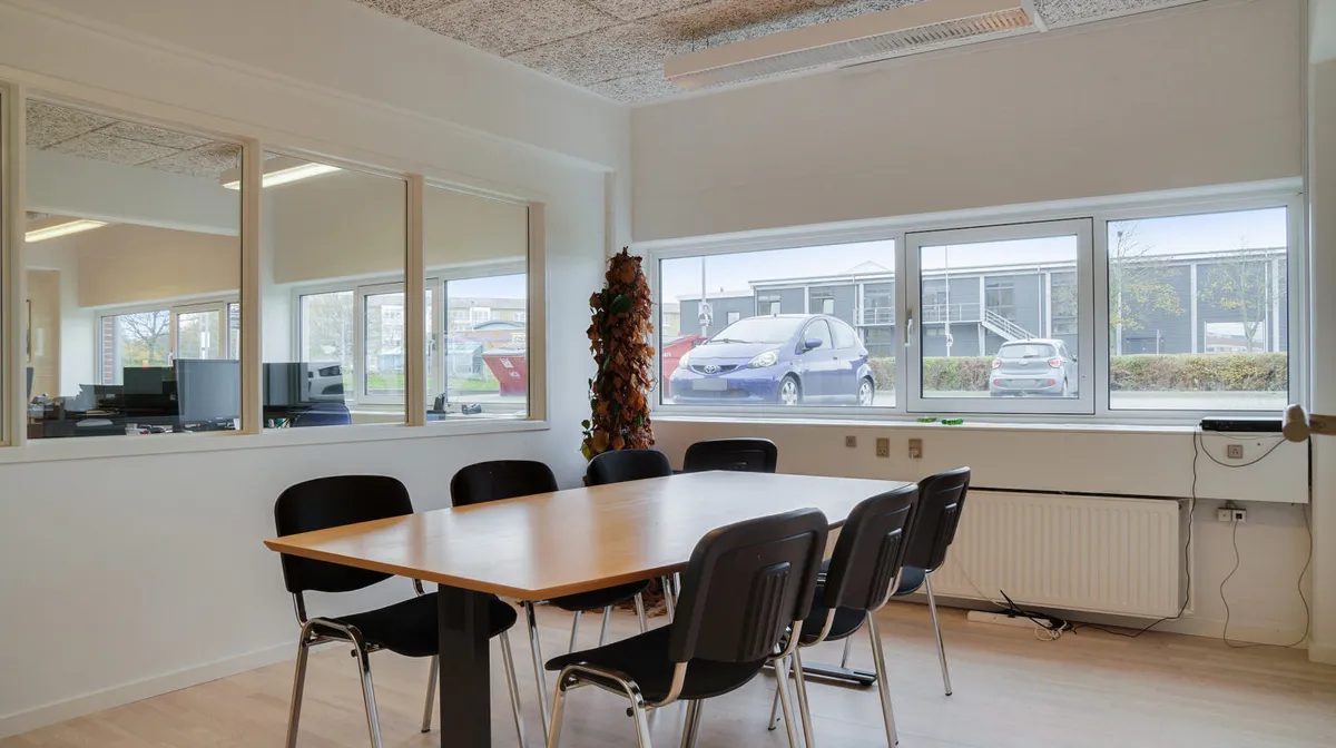 En kontorudlejning med en stor vinduesfront, en træbord og flere stole, et klimaanlæg og et landskabsbillede udenfor