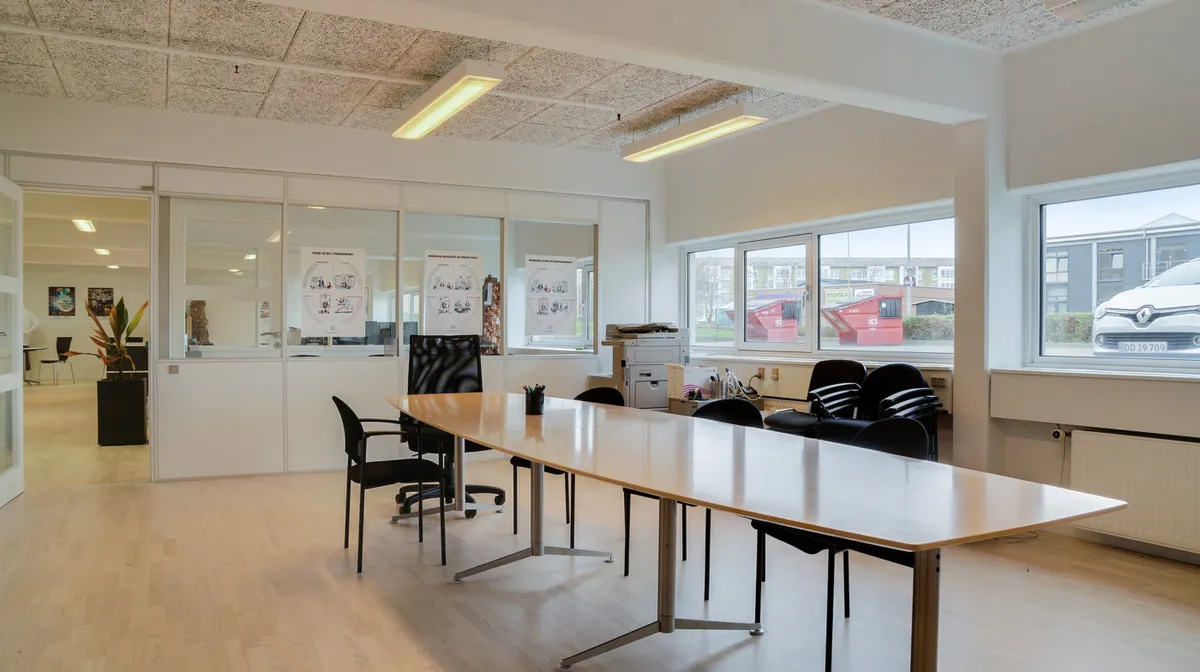 En stor kontorlokal med lysegulve, glasvægge, kontormøbler og en skrivemaskine i hjørnet samt en stor udsigt til gaden med