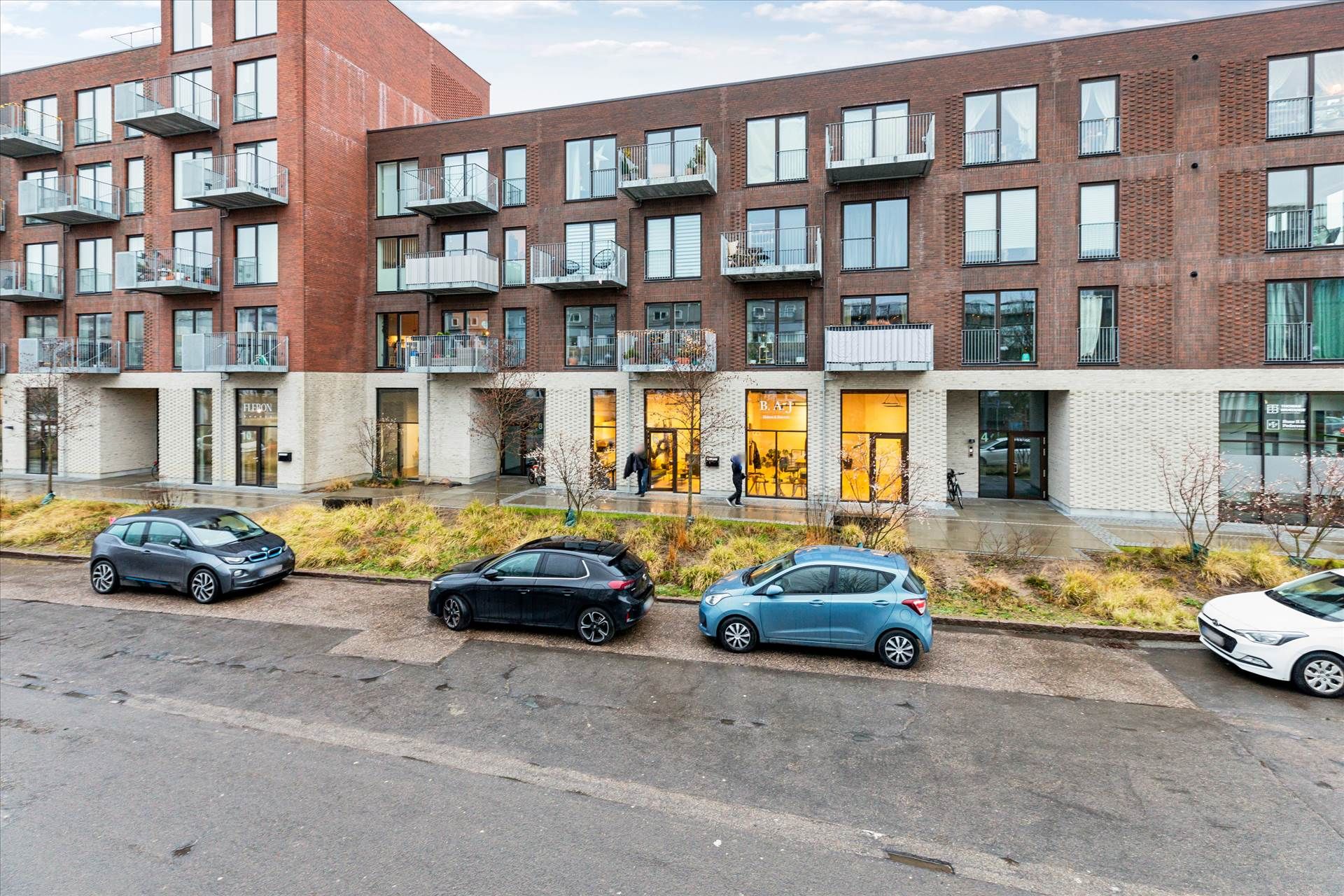 I billedet er der fem biler parkeret på gade, samt en gruppe mennesker går ind i en butik i et lejlighedskompleks