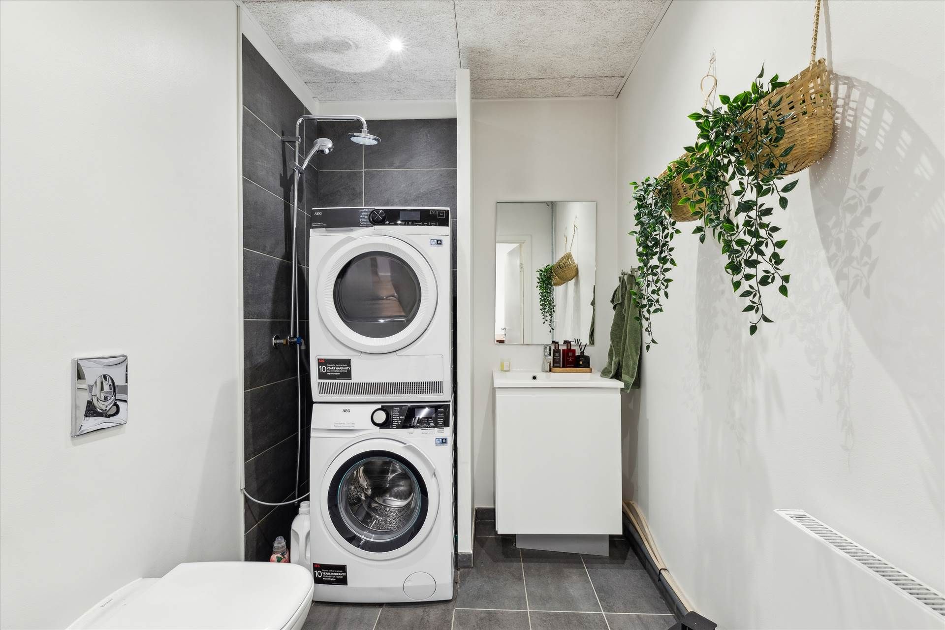 En moderne badeværelse med hvide vægge, to vaskemaskiner, et badekar, et spejl og en hængende plante
