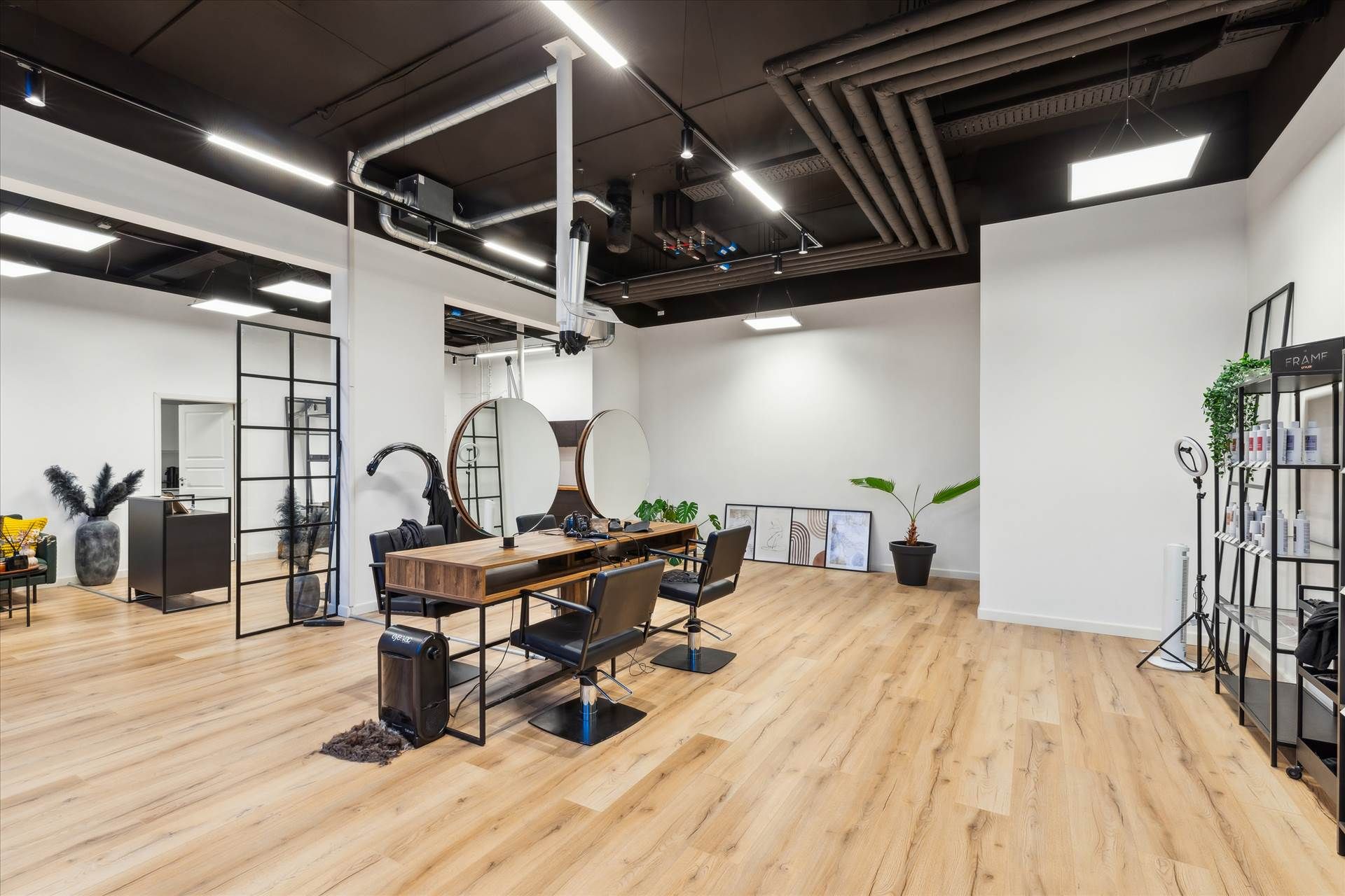 Store lyse stue med lysegul vinylgulv og højloft. Rundt bord med barberstole og spejle. Puder og plantepot.