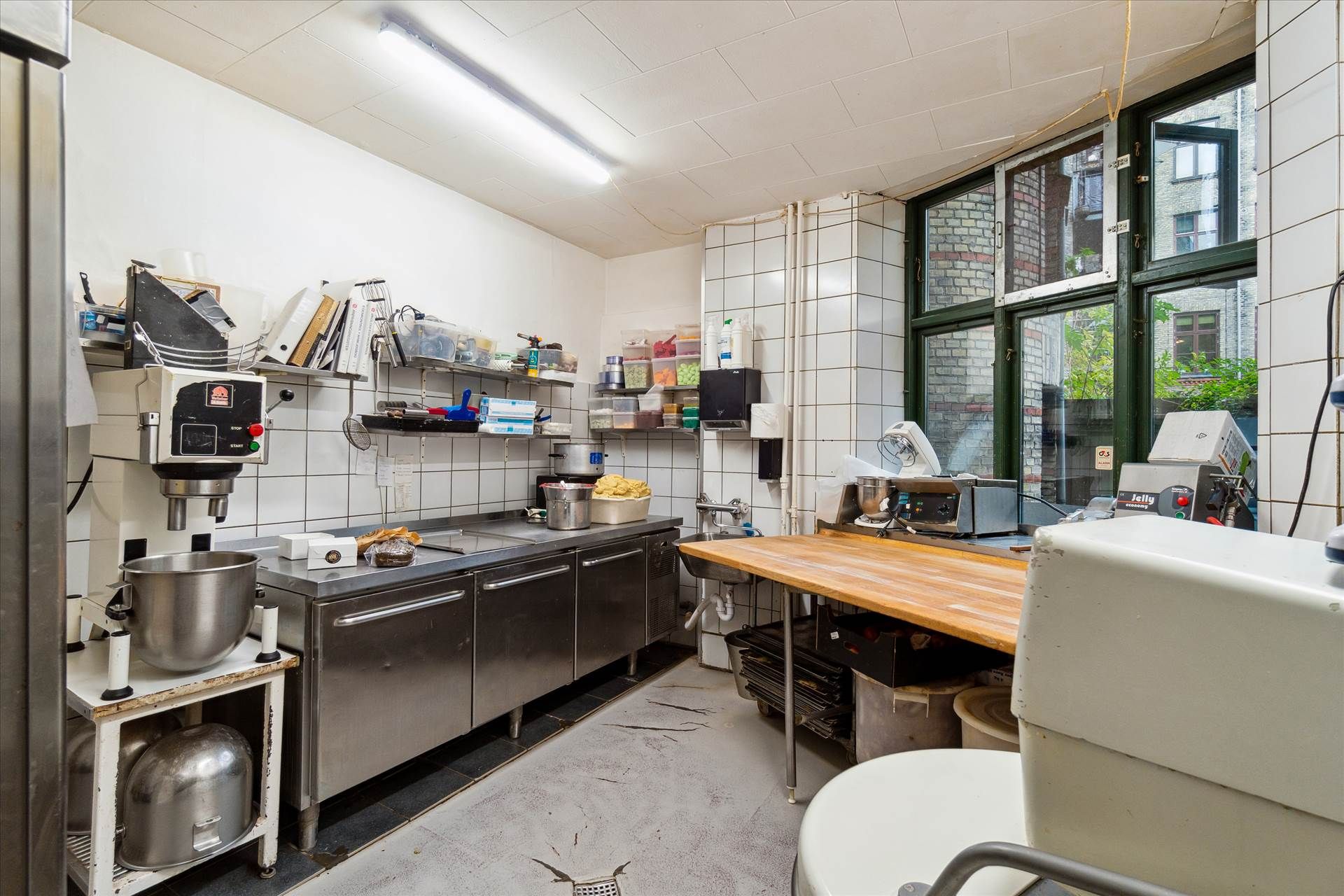 Bageri med rustfrit ståludstyr, brødrister og en stor vinduesvisning mod gaden. Lys, funktionel og udstyret med alle