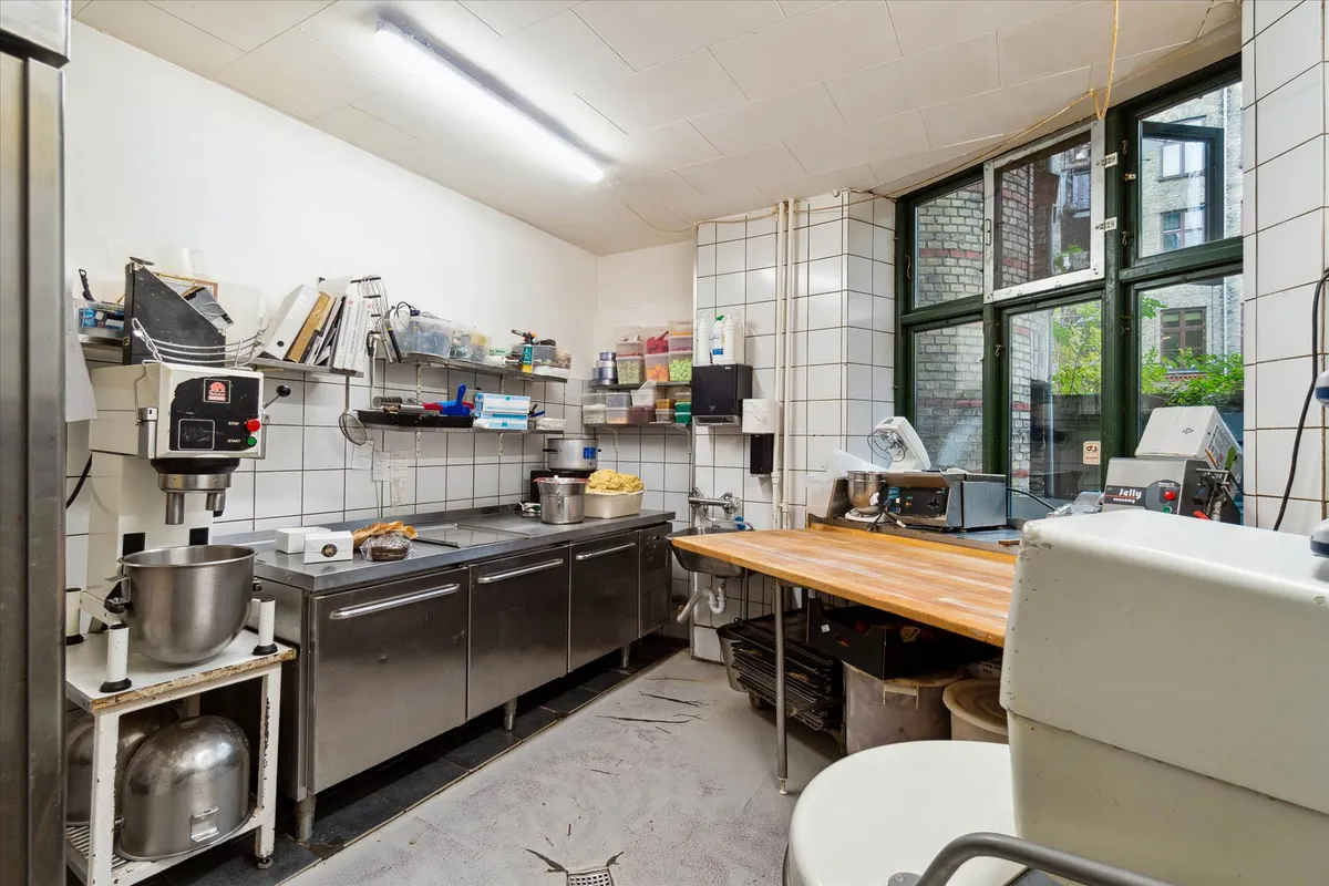 Bageri med rustfrit ståludstyr, brødrister og en stor vinduesvisning mod gaden. Lys, funktionel og udstyret med alle