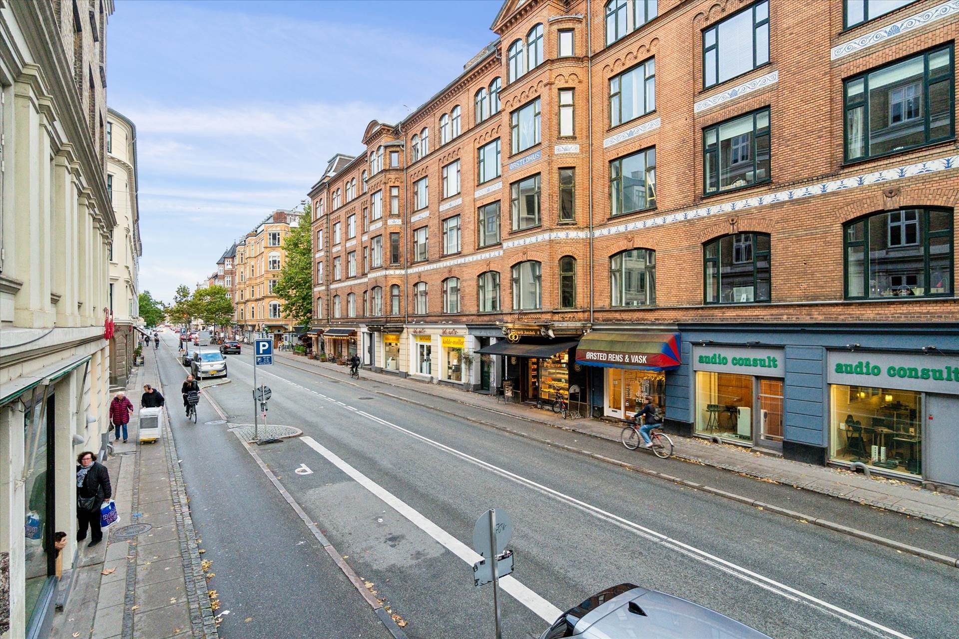En tom vej med butikker og mennesker på forsiden og biler og cyklister bagved. En parkeringsskilt og et skriftstykke er på