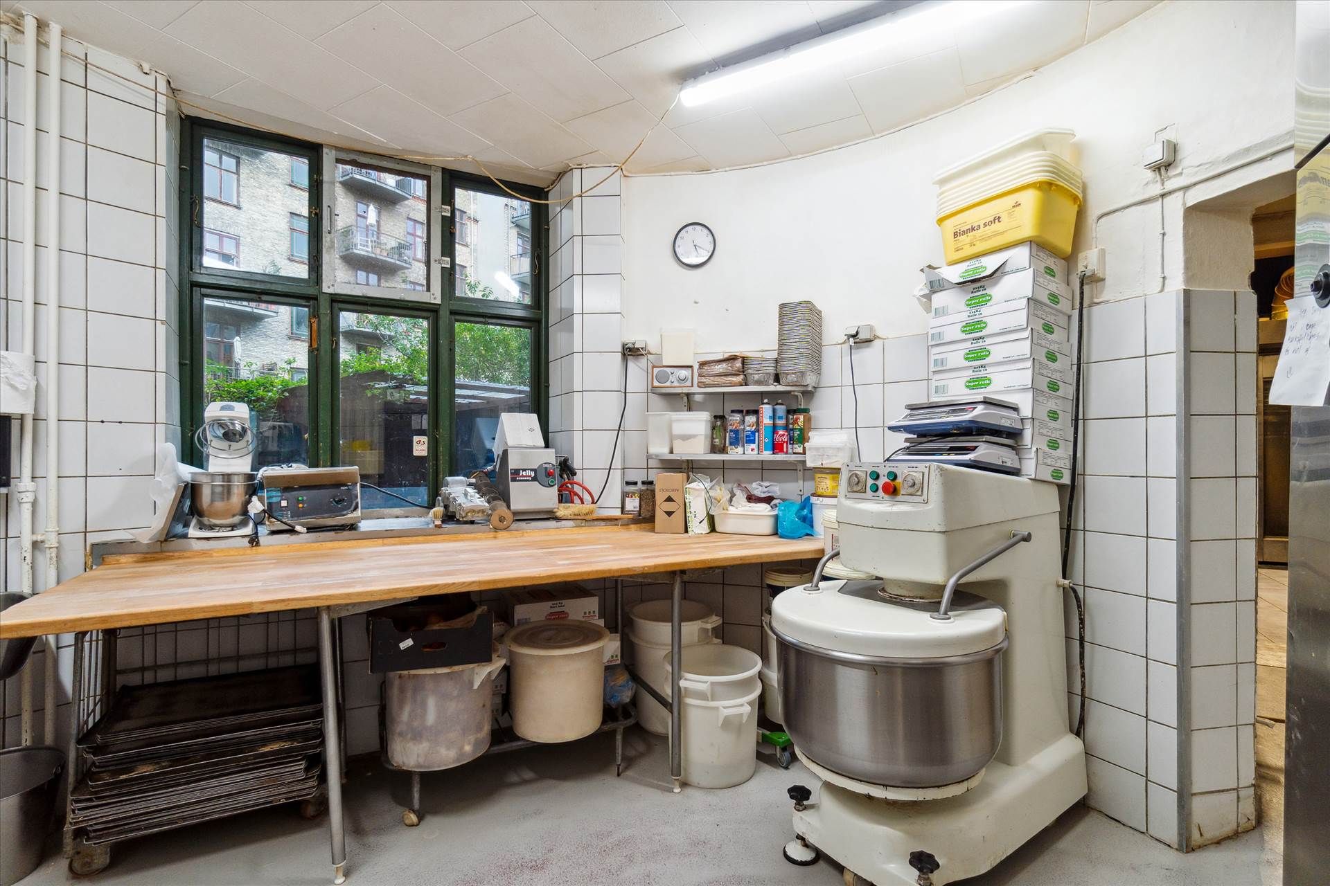 Bageri med køkkenbord og flere højtryksblandere samt bønner og ingredienser. Vindue med udsigt til bygning med altan.