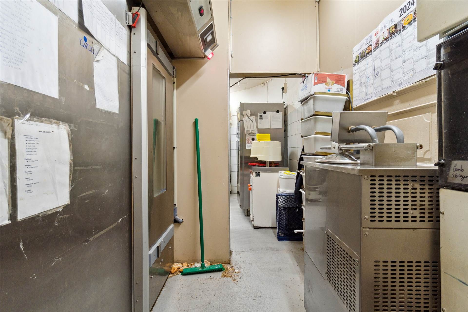 Moderne bageri med køleskab, fryser, komfur og ovn samt møbler og udstyr i et smal og langt rum med skrivepapir og kalender