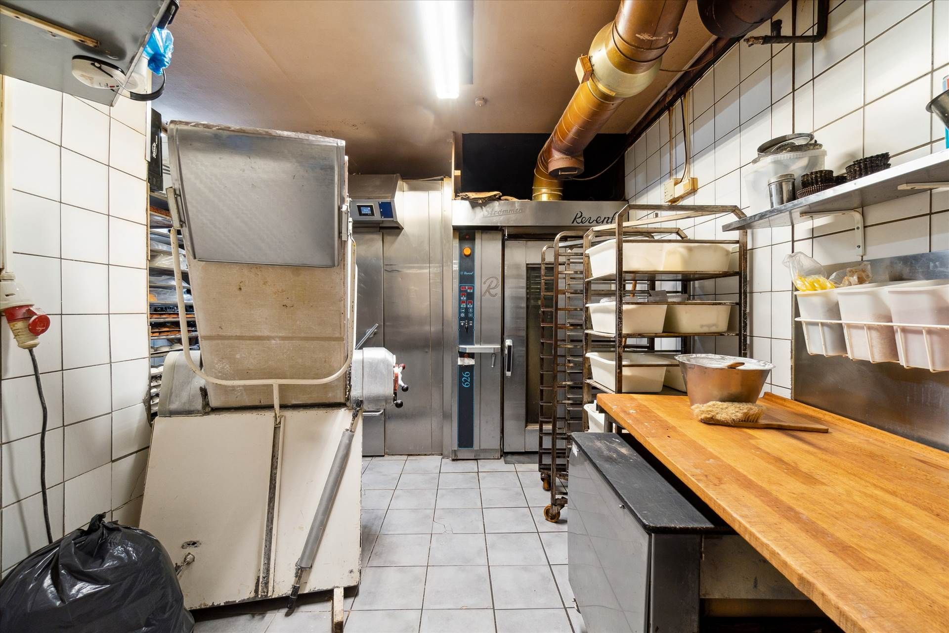 Bageri med ovn, vaskebøtte, træbord og flere stålhylder med bøtter. Lys og ren arbejdsplads med moderne udstyr.