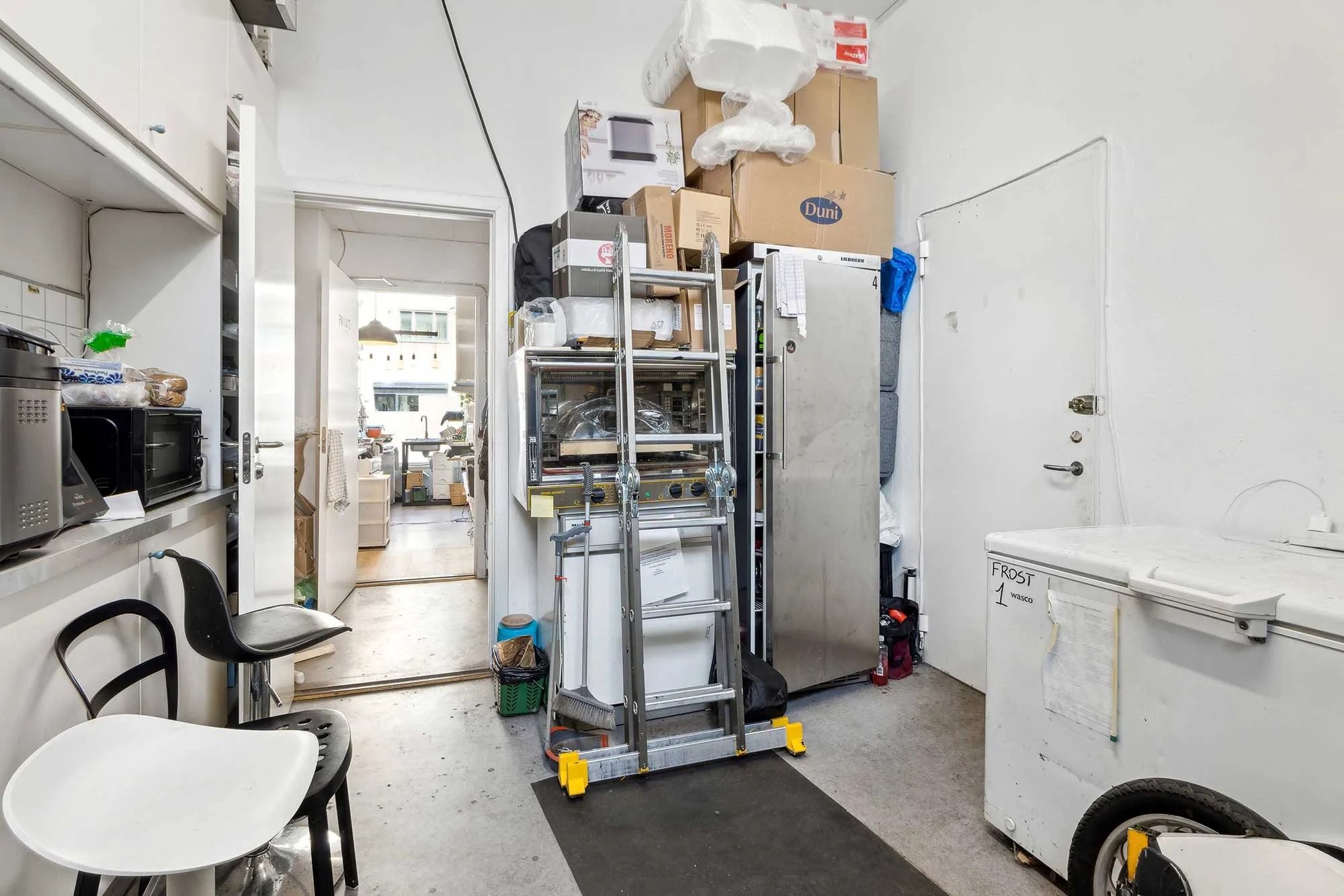 Lejlighed med et rummeligt køkken og opbevaringsrum, hvor flere køleskabe, ovne og skraldespande er opstillet