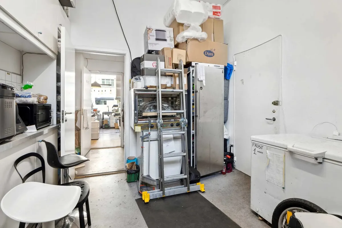 Lejlighed med et rummeligt køkken og opbevaringsrum, hvor flere køleskabe, ovne og skraldespande er opstillet