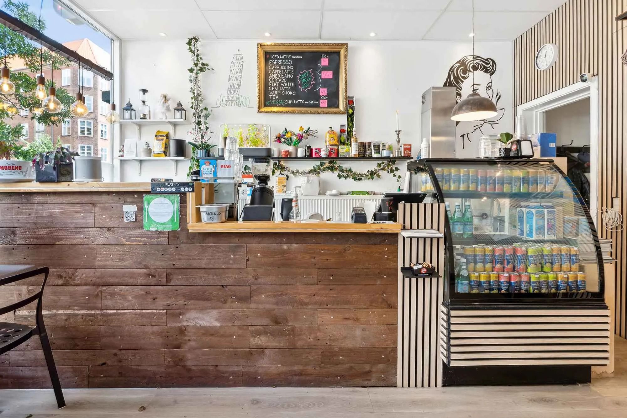 Kaffebar med træbar, menutavle, trævæg, glaskabinet med drikkevarer, loftslampe, blomster, plante og ure på væggen